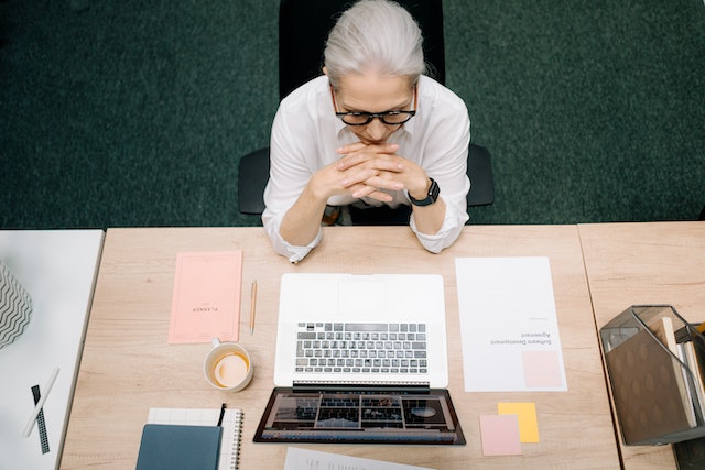 A grey haired woman sat at a desk with an open laptop