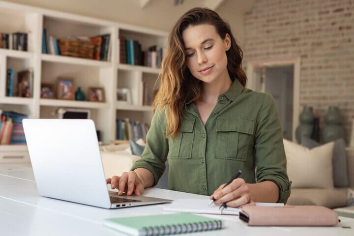 Young female sitting at desk with laptop and writing in notepad