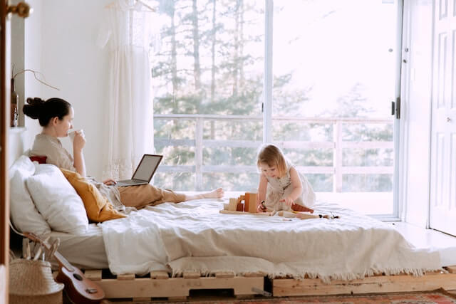 A woman studying on a bed with a laptop whilst her toddler plays next to her