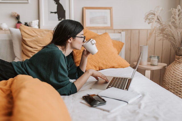 Woman sipping tea whilst studying on laptop on bed
